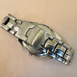 May include: A silver-toned wristwatch with a stainless steel band. The watch face is partially visible, and the band is closed. The watch is lying on a light brown surface.