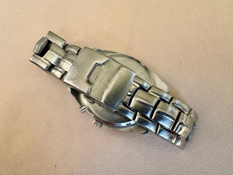 May include: A silver-toned wristwatch with a stainless steel band. The watch face is partially visible, and the band is closed. The watch is lying on a light brown surface.