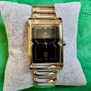 May include: A rectangular wristwatch with a two-tone silver and gold metal link band. The watch features a black dial with gold-tone hands, a date window displaying the number 17, and the Titan logo printed in gold at the top center position.
