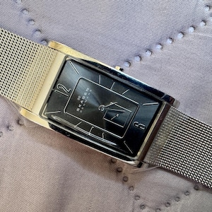 May include: A rectangular Skagen wristwatch featuring a black dial with silver-tone hands and markers, including the numerals 12 and 6. The watch has a polished silver-tone metal case and a fine silver-tone stainless steel mesh strap.