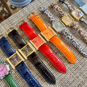May include: A collection of eight wristwatches arranged in a row. The watches feature various strap colors including green, blue, black, red, orange, gray leopard print, and brown leopard print, with square or round faces and gold-tone hardware.