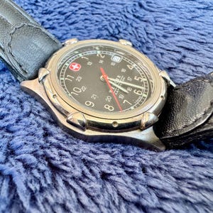 K&ouml;nnte beinhalten: Eine silberfarbene Armbanduhr mit schwarzem Zifferblatt, wei&szlig;en Ziffern und einem roten Sekundenzeiger. Die Uhr hat ein schwarzes Lederarmband und eine Datumsanzeige. Das Schweizer Kreuz-Logo ist sichtbar.