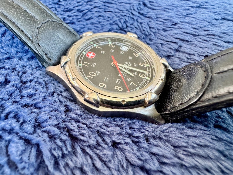 K&ouml;nnte beinhalten: Eine silberfarbene Armbanduhr mit schwarzem Zifferblatt, wei&szlig;en Ziffern und einem roten Sekundenzeiger. Die Uhr hat ein schwarzes Lederarmband und eine Datumsanzeige. Das Schweizer Kreuz-Logo ist sichtbar.