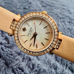 May include: A Kate Spade New York wristwatch with a cream-colored face and gold-tone hands. The watch has a gold bezel encrusted with crystals and a light brown leather band. The watch face features the Kate Spade logo and a mother-of-pearl effect.
