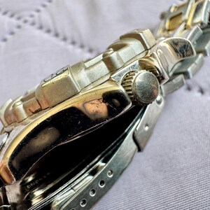 May include: A silver-tone metal wristwatch featuring a textured bezel and a matching link bracelet. The side of the watch case displays a circular crown with a ridged edge, while the polished metal surfaces reflect light across the watch body.