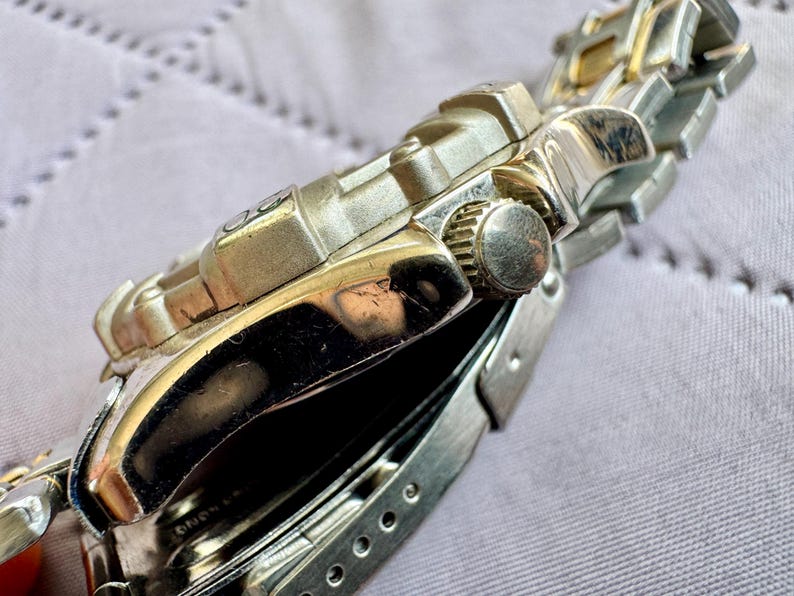 May include: A silver-tone metal wristwatch featuring a textured bezel and a matching link bracelet. The side of the watch case displays a circular crown with a ridged edge, while the polished metal surfaces reflect light across the watch body.