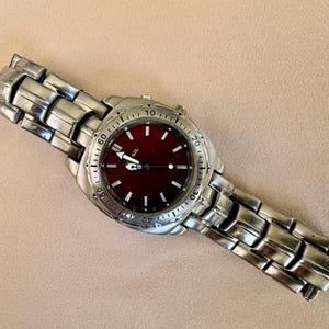 May include: A silver-toned wristwatch with a deep red face. The watch has a stainless steel band and a round dial with white markings. The watch hands are white, and the watch is set against a neutral background.