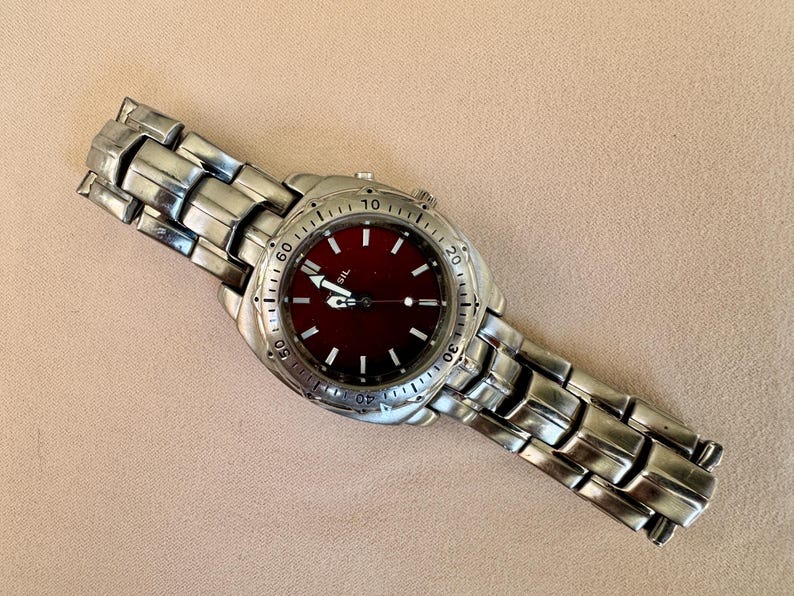 May include: A silver-toned wristwatch with a deep red face. The watch has a stainless steel band and a round dial with white markings. The watch hands are white, and the watch is set against a neutral background.