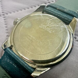 May include: A silver-tone Guess wristwatch with a teal leather strap. The back of the watch case features the Guess logo, 50 meters/165 feet water resistance, stainless steel back, model number U05168G6, and Japan movement text engraved.