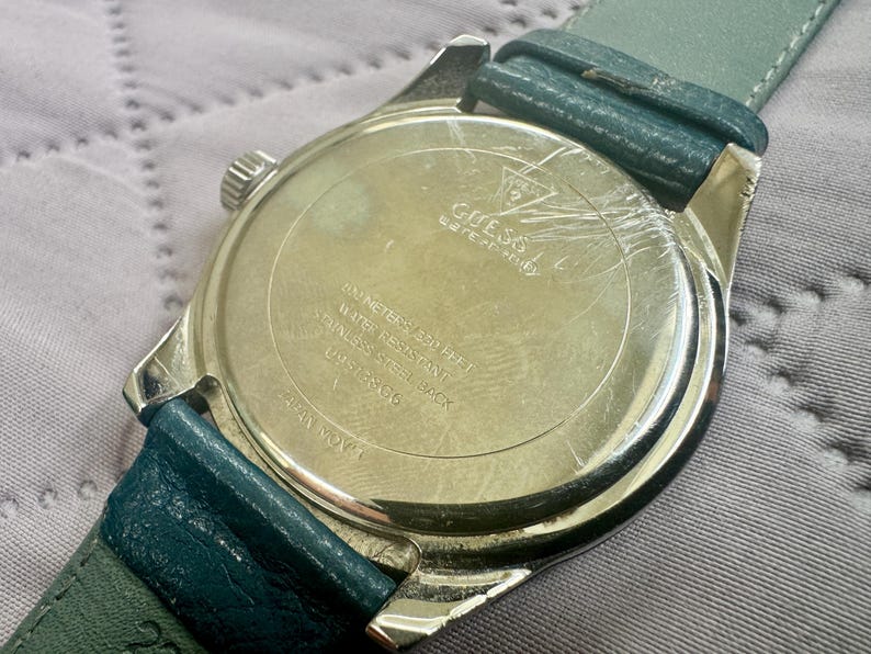 May include: A silver-tone Guess wristwatch with a teal leather strap. The back of the watch case features the Guess logo, 50 meters/165 feet water resistance, stainless steel back, model number U05168G6, and Japan movement text engraved.