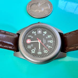 May include: A wristwatch with a black face, silver-tone case, and brown leather band. The watch face displays the time with white numerals and red accents. A silver coin is above the watch.