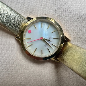 May include: Gold-toned Kate Spade watch with a pearlescent face, featuring a pink spade logo and gold hands. The watch has a gold leather band. The watch face is approximately 1 inch in diameter.