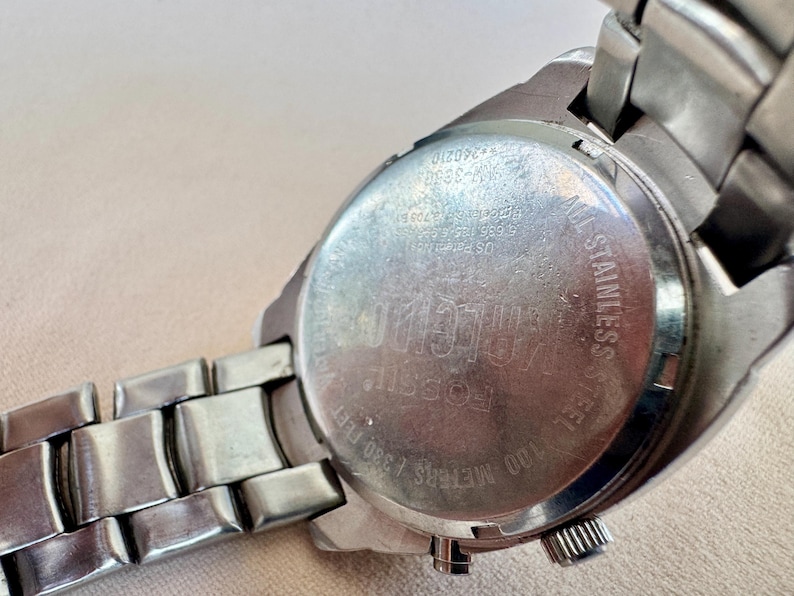 May include: Close-up of a silver-toned Fossil watch. The watch features a stainless steel back with the brand name and other details. The watch band is also silver-toned and made of metal links. The watch is water resistant up to 100 meters / 330 feet.