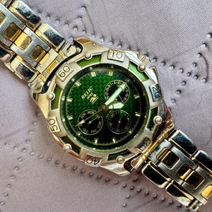 May include: A Relic Wet wristwatch featuring a green textured dial with three sub-dials and luminous hands. The watch has a silver and gold-tone metal link band and a rotating bezel with engraved numbers. The dial displays the brand name Relic Wet.