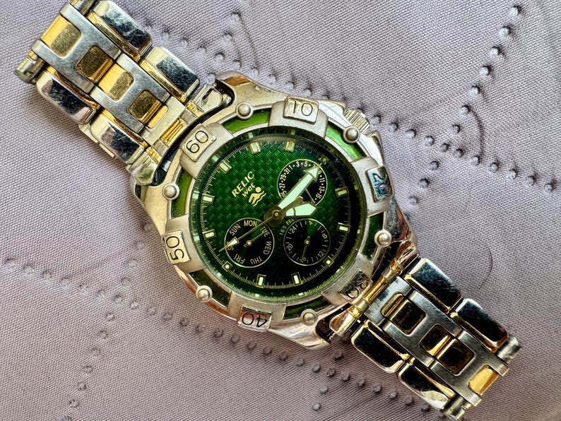 May include: A Relic Wet wristwatch featuring a green textured dial with three sub-dials and luminous hands. The watch has a silver and gold-tone metal link band and a rotating bezel with engraved numbers. The dial displays the brand name Relic Wet.