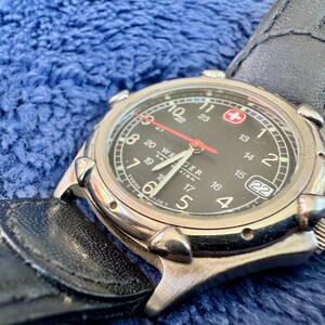 K&ouml;nnte beinhalten: Eine Wenger-Uhr mit schwarzem Zifferblatt, silberner L&uuml;nette und schwarzem Lederarmband. Die Uhr hat einen roten Sekundenzeiger, wei&szlig;e Ziffern und eine Datumsanzeige. Der Hintergrund ist blau strukturiert. Der Durchmesser des Zifferblatts betr&auml;gt ca. 3,8 cm.