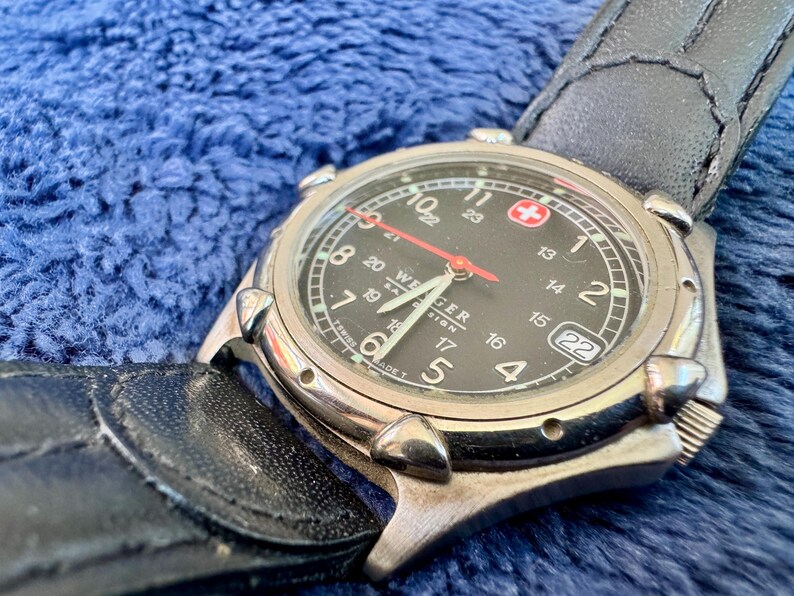 K&ouml;nnte beinhalten: Eine Wenger-Uhr mit schwarzem Zifferblatt, silberner L&uuml;nette und schwarzem Lederarmband. Die Uhr hat einen roten Sekundenzeiger, wei&szlig;e Ziffern und eine Datumsanzeige. Der Hintergrund ist blau strukturiert. Der Durchmesser des Zifferblatts betr&auml;gt ca. 3,8 cm.