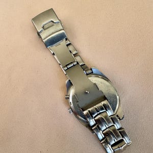 May include: A silver-toned wristwatch with a stainless steel band and clasp. The watch face is visible, and the band is partially extended. The watch is on a light brown surface.