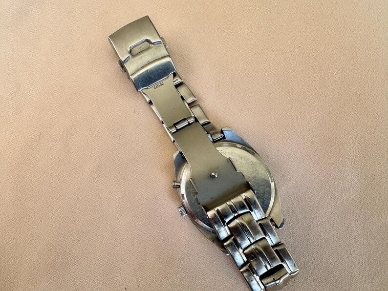 May include: A silver-toned wristwatch with a stainless steel band and clasp. The watch face is visible, and the band is partially extended. The watch is on a light brown surface.