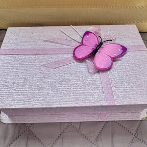May include: A rectangular light purple box with a textured surface, featuring a pink butterfly ornament attached to a light purple ribbon that wraps around the lid. The box has small metallic corner protectors on the front edges of the lid.