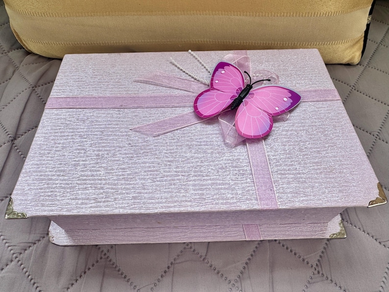 May include: A rectangular light purple box with a textured surface, featuring a pink butterfly ornament attached to a light purple ribbon that wraps around the lid. The box has small metallic corner protectors on the front edges of the lid.