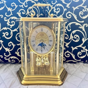 May include: A gold-tone mantel clock with a glass case and a handle on top. The clock face features Roman numerals, a gold filigree center, and a moon phase dial. A gold pendulum with four spheres hangs below the clock face within the glass enclosure.