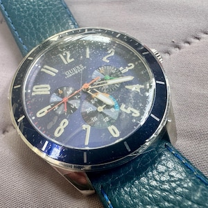 May include: A Guess wristwatch with a dark blue dial, silver-tone Arabic numerals, and three sub-dials. The watch features a silver-tone metal case, a dark blue rotating bezel, and a textured teal leather strap. The dial displays the brand name Guess.