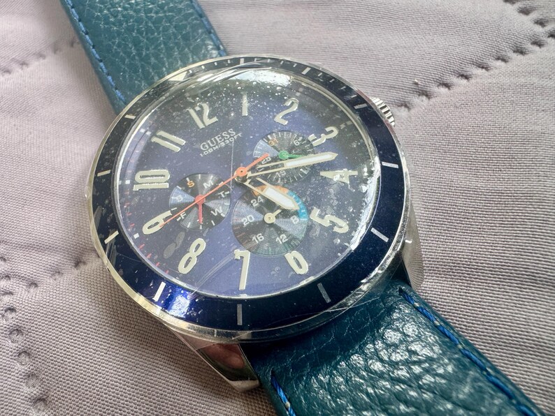 May include: A Guess wristwatch with a dark blue dial, silver-tone Arabic numerals, and three sub-dials. The watch features a silver-tone metal case, a dark blue rotating bezel, and a textured teal leather strap. The dial displays the brand name Guess.