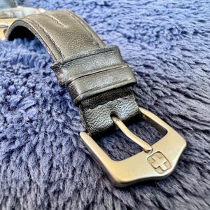 K&ouml;nnte beinhalten: Nahaufnahme eines schwarzen Leder-Armbands mit silberner Schnalle. Die Schnalle hat ein Kreuzemblem. Das Armband liegt auf einer blauen, strukturierten Oberfl&auml;che.