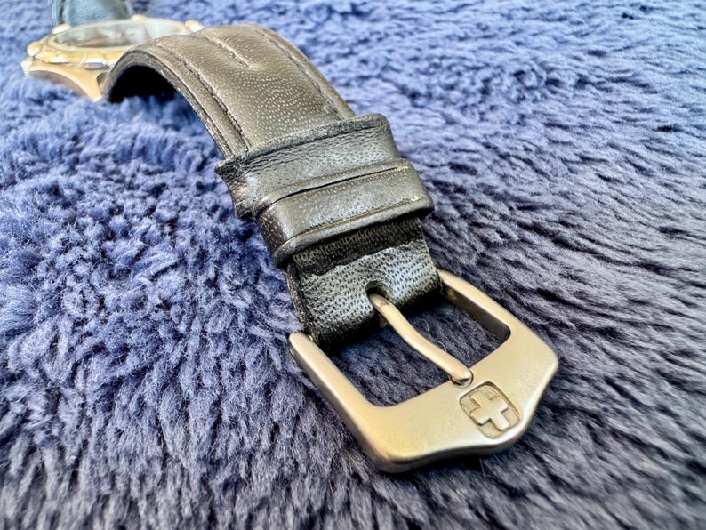 K&ouml;nnte beinhalten: Nahaufnahme eines schwarzen Leder-Armbands mit silberner Schnalle. Die Schnalle hat ein Kreuzemblem. Das Armband liegt auf einer blauen, strukturierten Oberfl&auml;che.