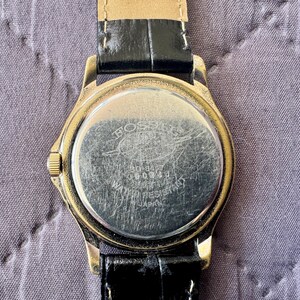 K&ouml;nnte beinhalten: Eine goldfarbene Armbanduhr mit schwarzem Lederarmband. Der runde Geh&auml;useboden aus silberfarbenem Metall weist sichtbare Kratzer auf und tr&auml;gt die Gravur Fossil, 50-8437, 060004, Water Resistant und Japan. Eine kleine goldfarbene Krone befindet sich seitlich.