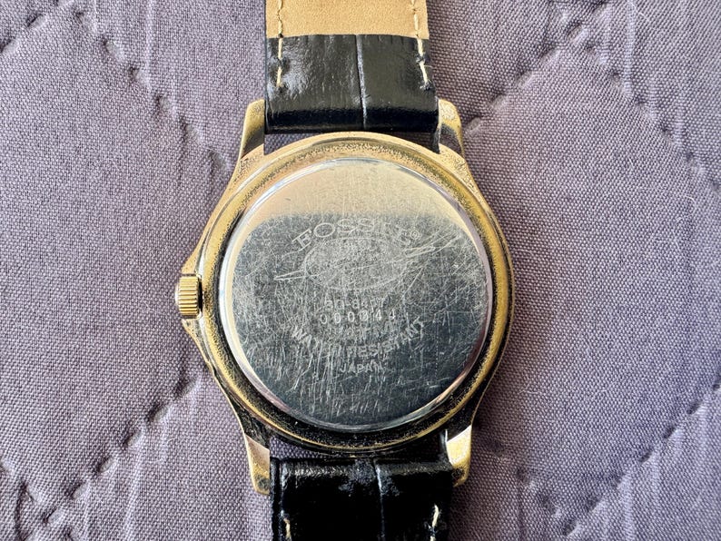 K&ouml;nnte beinhalten: Eine goldfarbene Armbanduhr mit schwarzem Lederarmband. Der runde Geh&auml;useboden aus silberfarbenem Metall weist sichtbare Kratzer auf und tr&auml;gt die Gravur Fossil, 50-8437, 060004, Water Resistant und Japan. Eine kleine goldfarbene Krone befindet sich seitlich.
