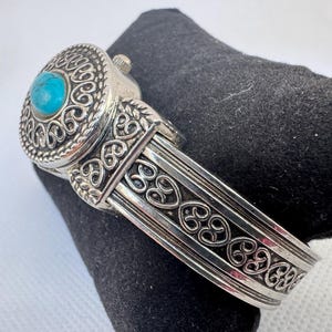 Puede incluir: Reloj de pulsera plateado con un dise&ntilde;o ornamentado. La esfera del reloj presenta una piedra turquesa redonda engastada en un bisel decorativo. La pulsera est&aacute; detallada con patrones de volutas. El reloj est&aacute; sobre una superficie negra.