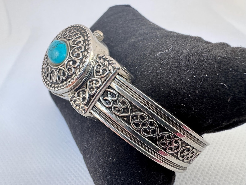 Puede incluir: Reloj de pulsera plateado con un dise&ntilde;o ornamentado. La esfera del reloj presenta una piedra turquesa redonda engastada en un bisel decorativo. La pulsera est&aacute; detallada con patrones de volutas. El reloj est&aacute; sobre una superficie negra.