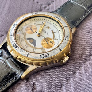 K&ouml;nnte beinhalten: Eine Fossil Armbanduhr mit wei&szlig;em Zifferblatt, goldfarbenen Akzenten, einer Mondphasenanzeige und zwei Hilfszifferbl&auml;ttern. Die Uhr hat eine gold- und silberfarbene Metalll&uuml;nette mit eingravierten Zahlen und ein schwarzes, strukturiertes Lederarmband.