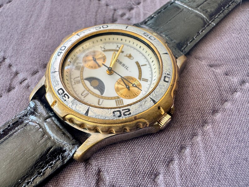 K&ouml;nnte beinhalten: Eine Fossil Armbanduhr mit wei&szlig;em Zifferblatt, goldfarbenen Akzenten, einer Mondphasenanzeige und zwei Hilfszifferbl&auml;ttern. Die Uhr hat eine gold- und silberfarbene Metalll&uuml;nette mit eingravierten Zahlen und ein schwarzes, strukturiertes Lederarmband.