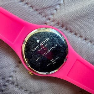 May include: A bright pink Kate Spade watch with a gold-colored bezel and a black watch face. The watch band is made of a matching pink material. The watch face reads "Kate Spade New York" and "Live Colorfully".