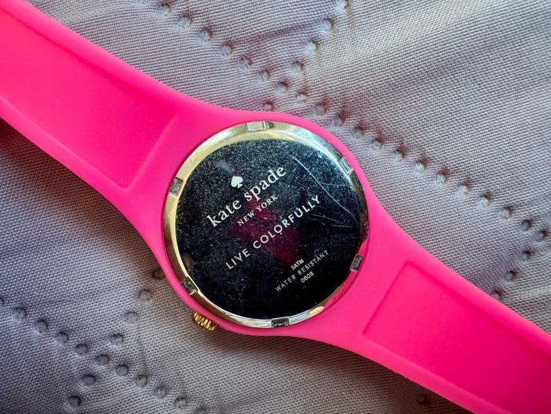 May include: A bright pink Kate Spade watch with a gold-colored bezel and a black watch face. The watch band is made of a matching pink material. The watch face reads "Kate Spade New York" and "Live Colorfully".