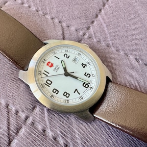 K&ouml;nnte beinhalten: Eine Swiss Army Armbanduhr mit rundem Edelstahlgeh&auml;use, wei&szlig;em Zifferblatt mit schwarzen arabischen Ziffern, Datumsanzeige auf der Drei-Uhr-Position und braunem Lederarmband. Das Zifferblatt zeigt das Swiss Army Logo und den Schriftzug.