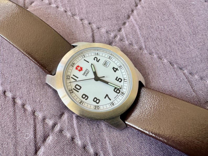 K&ouml;nnte beinhalten: Eine Swiss Army Armbanduhr mit rundem Edelstahlgeh&auml;use, wei&szlig;em Zifferblatt mit schwarzen arabischen Ziffern, Datumsanzeige auf der Drei-Uhr-Position und braunem Lederarmband. Das Zifferblatt zeigt das Swiss Army Logo und den Schriftzug.