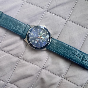 May include: A wristwatch featuring a round silver-tone case with a dark blue dial and a textured teal leather strap. The dial displays silver-tone Arabic numerals, three sub-dials, and a silver-tone metal buckle closure on the strap.
