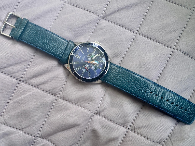 May include: A wristwatch featuring a round silver-tone case with a dark blue dial and a textured teal leather strap. The dial displays silver-tone Arabic numerals, three sub-dials, and a silver-tone metal buckle closure on the strap.