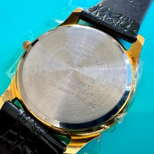 Peut inclure: Gros plan d'une montre dor&eacute;e avec un bracelet en cuir noir. Le dos de la montre est argent&eacute; avec le texte "BASE METAL BEZEL, STAINLESS STEEL BACK, SWISS MOVT PARTS, WATER RESISTANT 100FT." La montre est &eacute;tanche &agrave; 30,48 m&egrave;tres.
