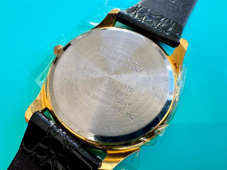 Peut inclure: Gros plan d'une montre dor&eacute;e avec un bracelet en cuir noir. Le dos de la montre est argent&eacute; avec le texte "BASE METAL BEZEL, STAINLESS STEEL BACK, SWISS MOVT PARTS, WATER RESISTANT 100FT." La montre est &eacute;tanche &agrave; 30,48 m&egrave;tres.