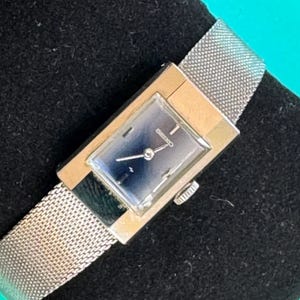 May include: A vintage Seiko watch with a rectangular face and a mesh metal band. The watch face is a deep blue color with silver hands and hour markers. The watch is set against a black background.