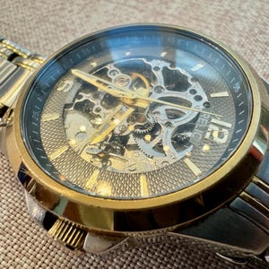 May include: A Relic brand wristwatch with a gold-tone bezel and a silver-tone band. The watch face is black with gold-tone hands and hour markers. The watch features a skeleton design, revealing the inner workings.