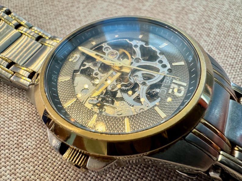 May include: A Relic brand wristwatch with a gold-tone bezel and a silver-tone band. The watch face is black with gold-tone hands and hour markers. The watch features a skeleton design, revealing the inner workings.