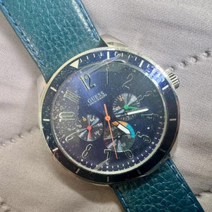 May include: A Guess wristwatch with a dark blue dial, silver-tone numerals, and three sub-dials. The watch features a textured teal leather strap, a silver-tone metal case, and a cracked glass face. The dial displays the text GUESS 100M/330FT.