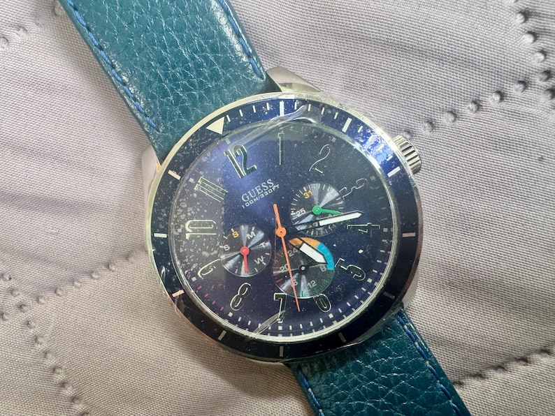 May include: A Guess wristwatch with a dark blue dial, silver-tone numerals, and three sub-dials. The watch features a textured teal leather strap, a silver-tone metal case, and a cracked glass face. The dial displays the text GUESS 100M/330FT.