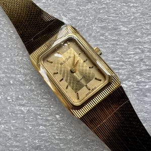 May include: A gold-tone Bulova quartz watch with a rectangular face and a textured gold dial. The watch features a matching gold-tone metal mesh strap with a fine linear pattern. The dial displays the Bulova logo and the word Quartz.